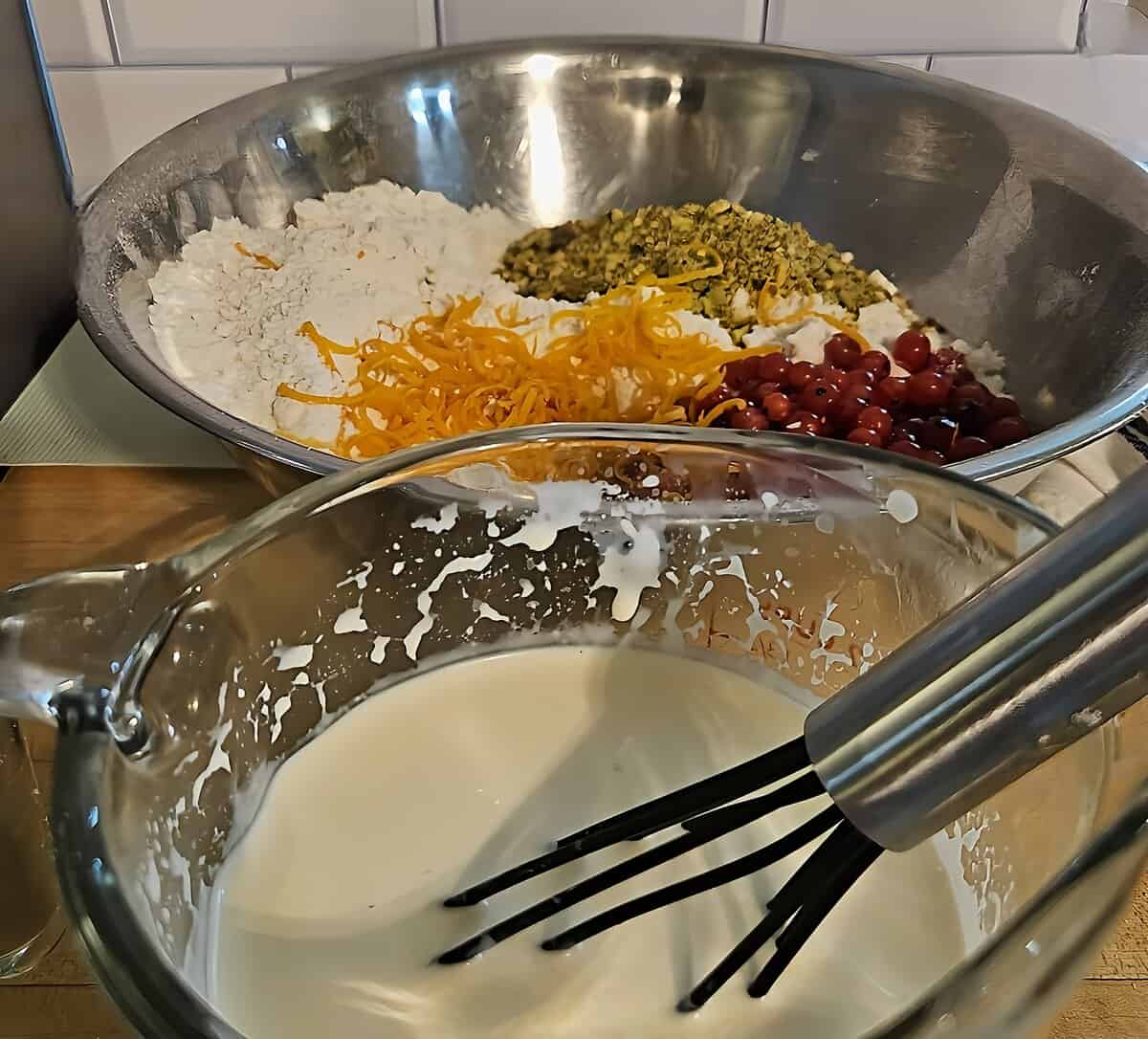 dry and wet ingredients for scone dough shown separately. half and half in measuring cup with a whip in it, flour with butter cut into it with currants, orange zest, and pistachios resting on top in a mixing bowl