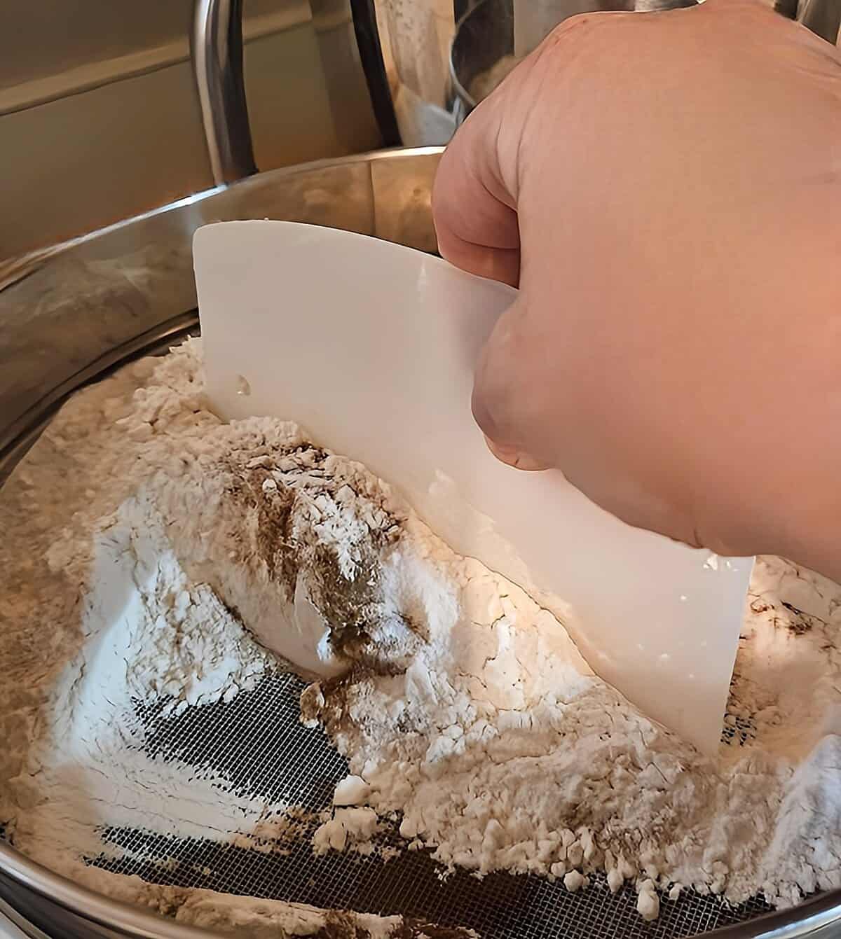 dry ingredients being pushed through a drum sieve with a plastic dough scraper