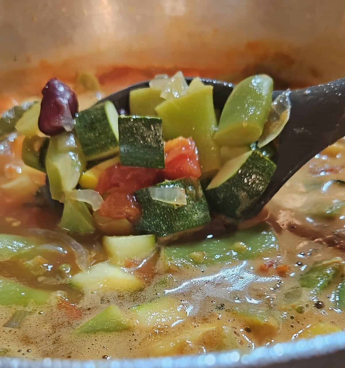 minestrone soup being simmered in a saucepan, with spoon lifting out the vegetables and stirring