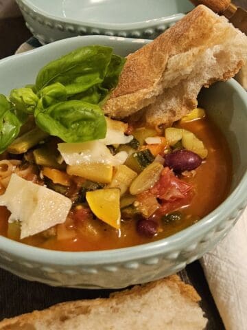 finished minestrone in a bowl for serving with chunks of parmesan cheese, a fresh basil top, and a chunk of crusty baguette