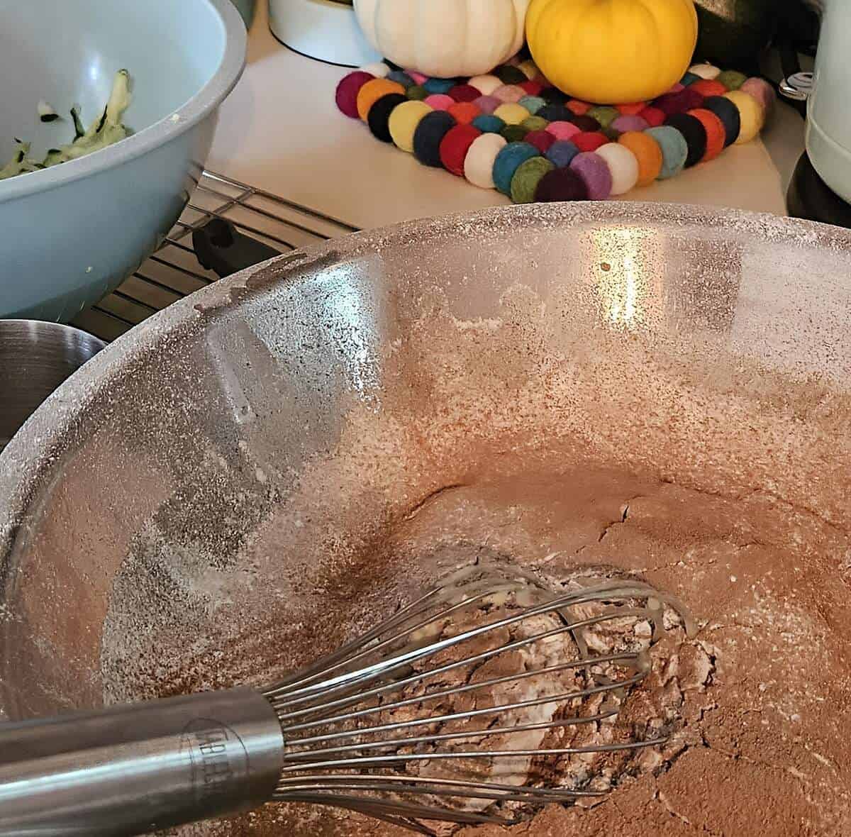 zucchini brownie batter being mixed in a bowl with a wire whip, shredded zucchini in background