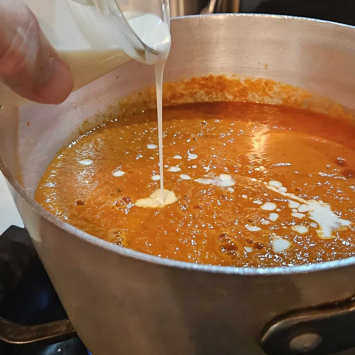 tomato soup in a saucepan with heavy cream being poured in.