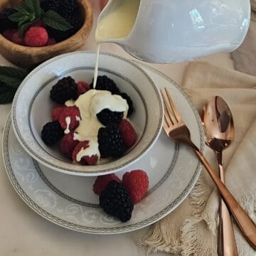 crème anglaise sauce being poured from a small porcelain pitcher on top of berries in a bowl