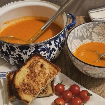 finished tomato soup, in a enamel crock with a ladle, in a bowl with a spoon, a grilled cheese sandwich, and fresh cherry tomatoes.