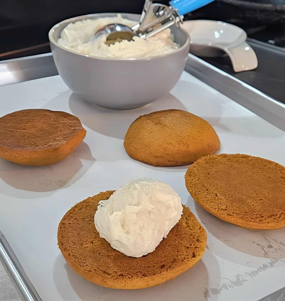 assembly of pumpkin whoopie pies; one cake circle with a scoop f filling on top, three cakes set aside naked, bowl of filling with a scoop in it, ready to scoop more
