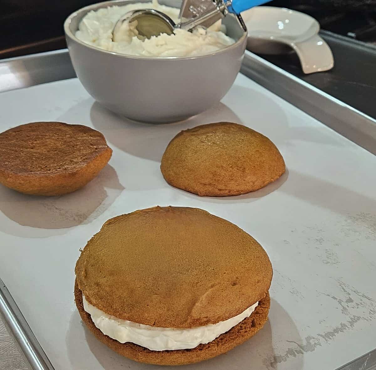 assembly of pumpkin whoopie pies, fully assembled cakes with cream cheese filling in between them, two cakes laying aside with no filling, filling in a bowl with a scoop in it ready to be used for filling.
