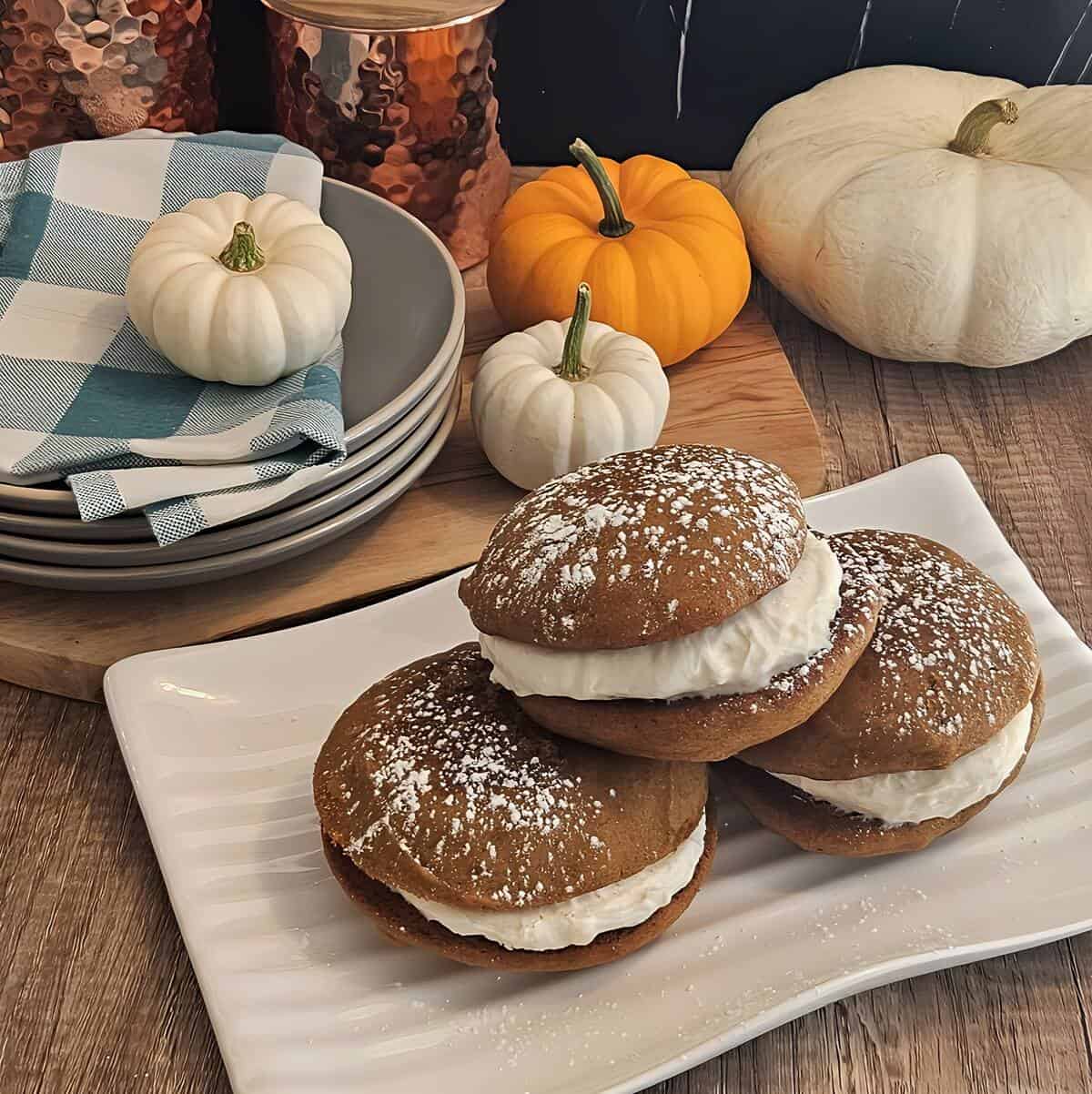 finished whoopie pies, small round cakes with cream cheese filling in between, on plate stacked on top of each other, topped with powdered sugar, fall decorations.