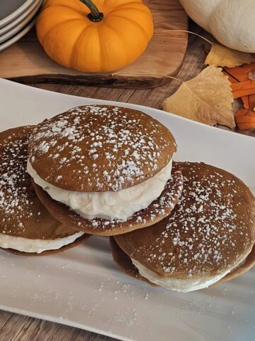 finished whoopie pies, small round cakes with cream cheese filling in between, on plate stacked on top of each other, topped with powdered sugar, fall decorations.