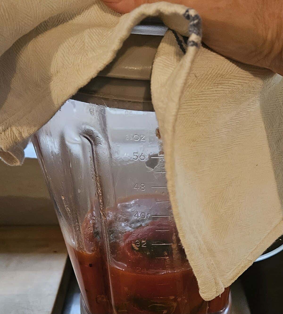 blender loaded with unprocessed tomato soup, a hand covering the lid with a dry towel.