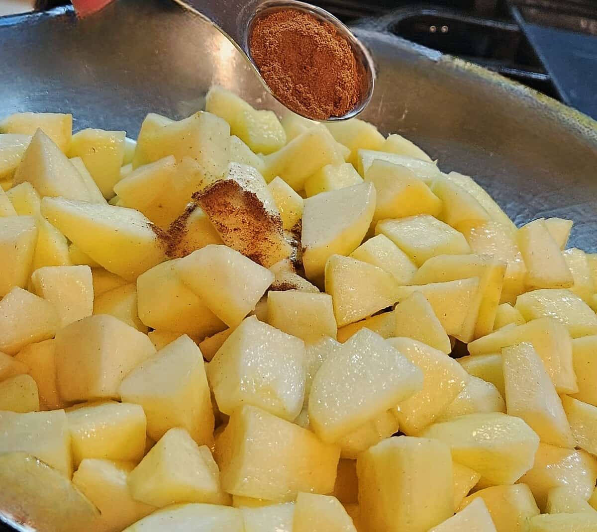 chunks of apple in skillet with spices being sprinkled in with a tablespoon