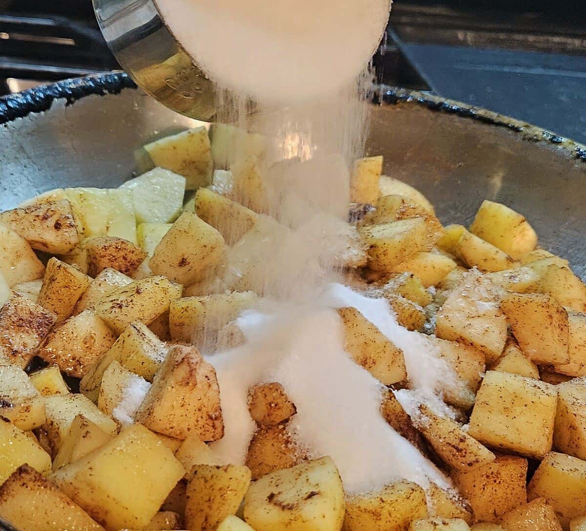 granulated sugar being poured into a skillet of spiced and cooking apple chunks.