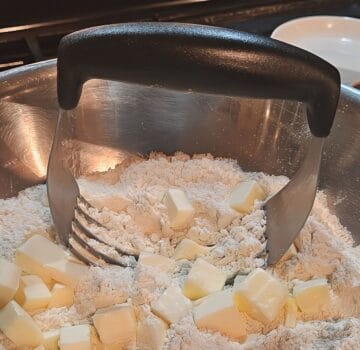 dry ingredients for biscuit topping with chunks of butter in it, pastry cutter resting in pile inside of mixing bowl.