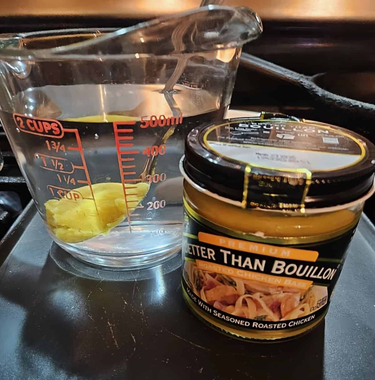 measuring cup filled with water, with a spoonful of chicken bouillon submerged in it. jar of bouillon in foreground.
