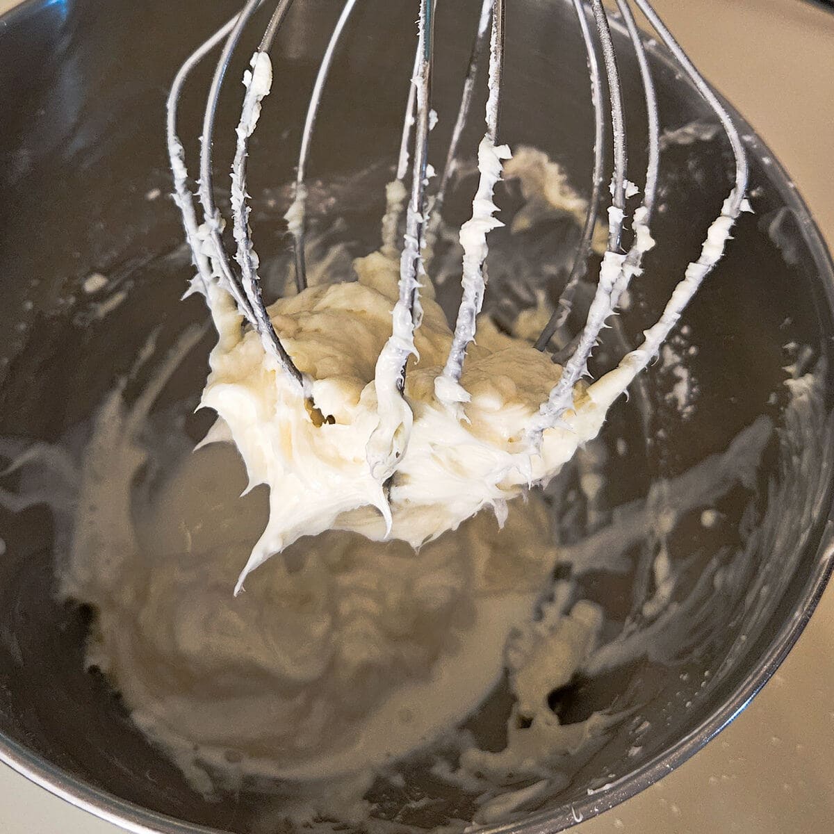 cream cheese icing for cinnamon rolls in stand mixer, wire whip attachment with some of it stuck to it, showing creamy consistency.