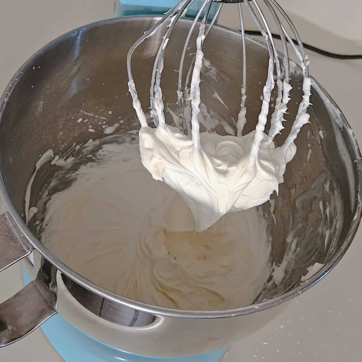 cream cheese icing in a stand mixer, clinging to the whip attachment showing finished fluffy texture.