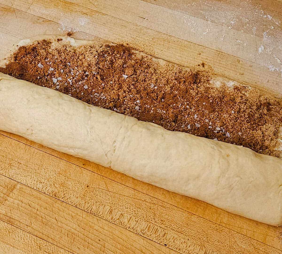 cinnamon roll dough with butter, brown sugar, cinnamon and sea salt filling being rolled into a log.