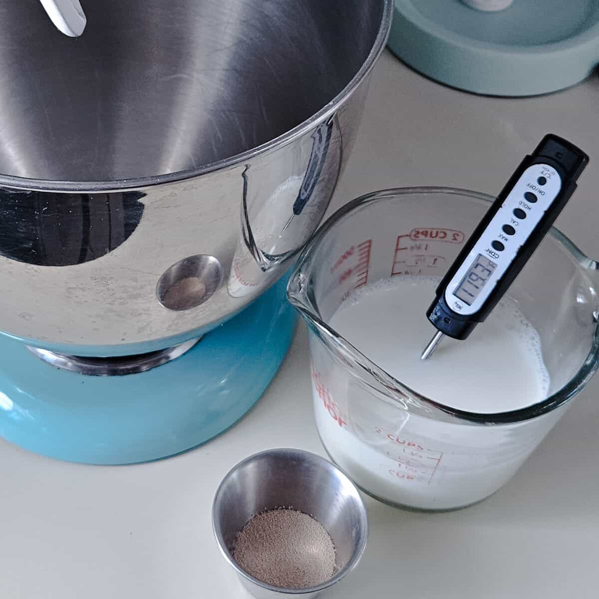 measuring cup with milk in it, measuring to 119.3 degrees F with a digital thermometer, yeast granules to the side in a metal ramekin, stand mixer waiting in the background.