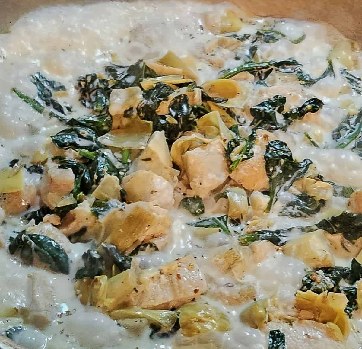 chicken, spinach, and artichoke hearts in a skillet with cream bubbling around it.