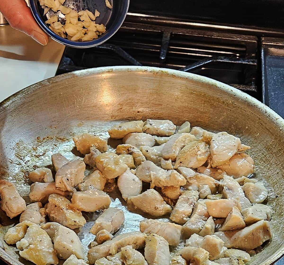 seared chicken breast pieces in a skillet with a ramekin of minced garlic being dropped in.