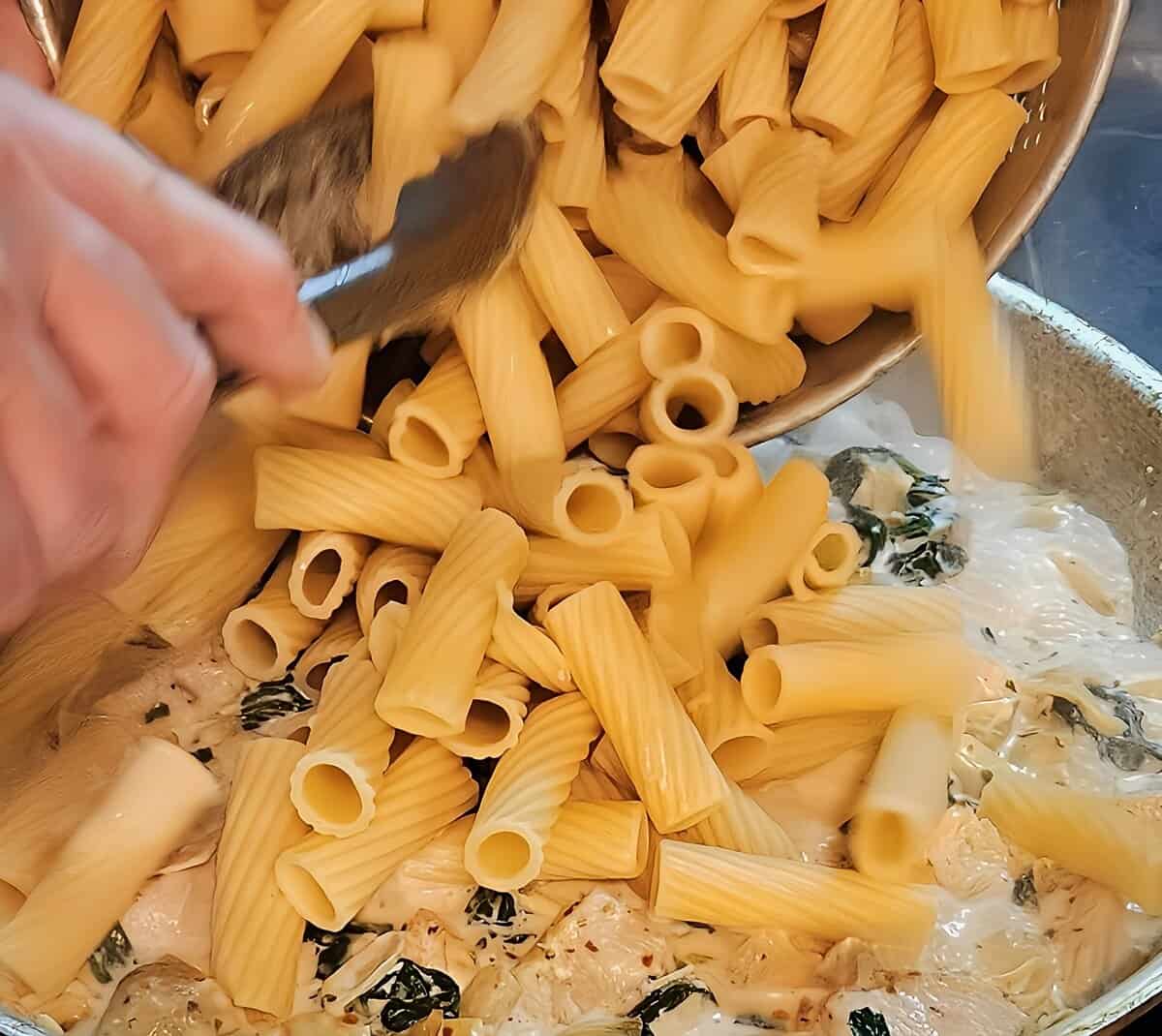 cooked rigatoni pasta being poured into a skillet containing chicken, spinach, and artichokes cooking in a cream sauce.