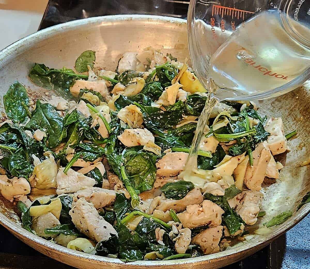chicken, spinach, and artichoke hearts in a skillet with white wine being poured in.