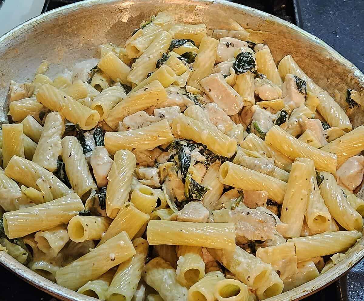 spinach chicken artichoke pasta finished in a skillet. rigatoni pasta, spinach, chicken breast chunks, and artichoke heats coated in thick creamy sauce.