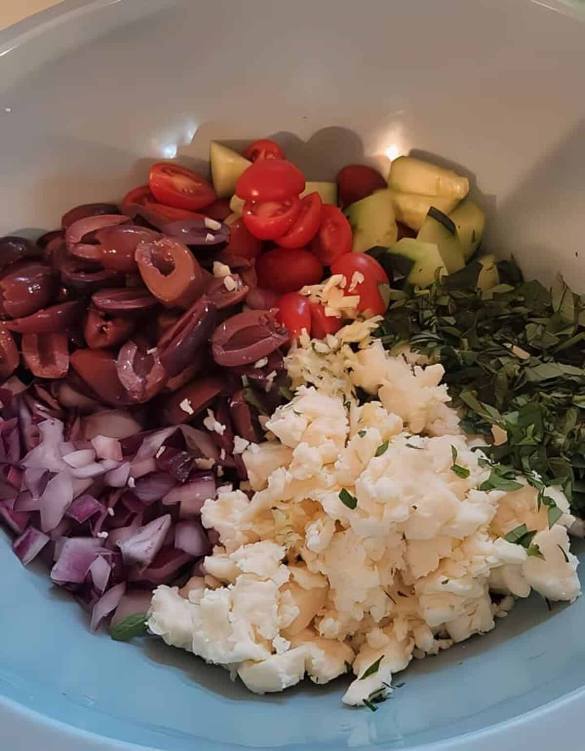 chopped ingredients for greek orzo salad. oregano, cucumber, tomato, olives, red onion, and feta cheese.