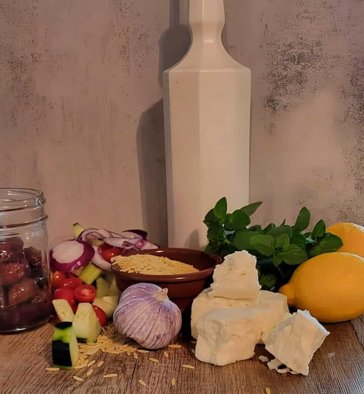 ingredients for greek orzo pasta salad, olives, tomatoes. onion, cucumber, raw orzo, garlic bulb, feta, oregano, lemons, bottle of olive oil