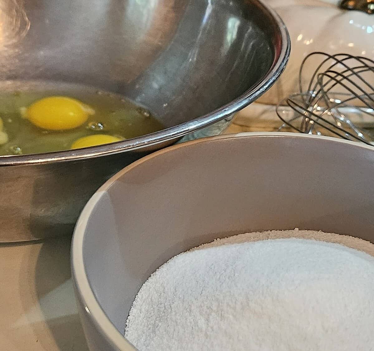 mixing bowl with eggs cracked in it, bowl of sugar, wire whip