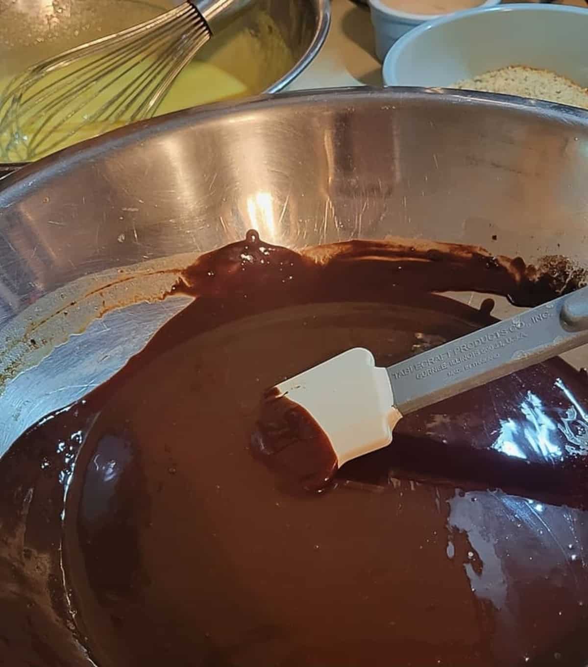 melted chocolate in metal mixing bowl with rubber spatula, egg yolk mixture in bowl with wire whip in background