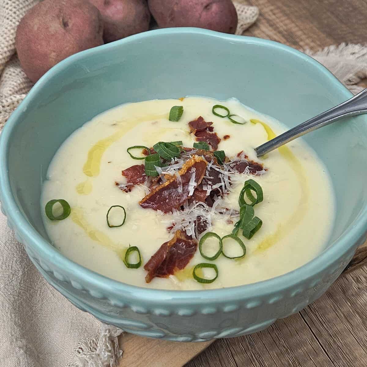 the potato and shallot soup with crispy prosciutto in a bowl garnished with crispy prosciutto, green onion, parmesan cheese, and the oil from roasting the shallots and garlic closeup