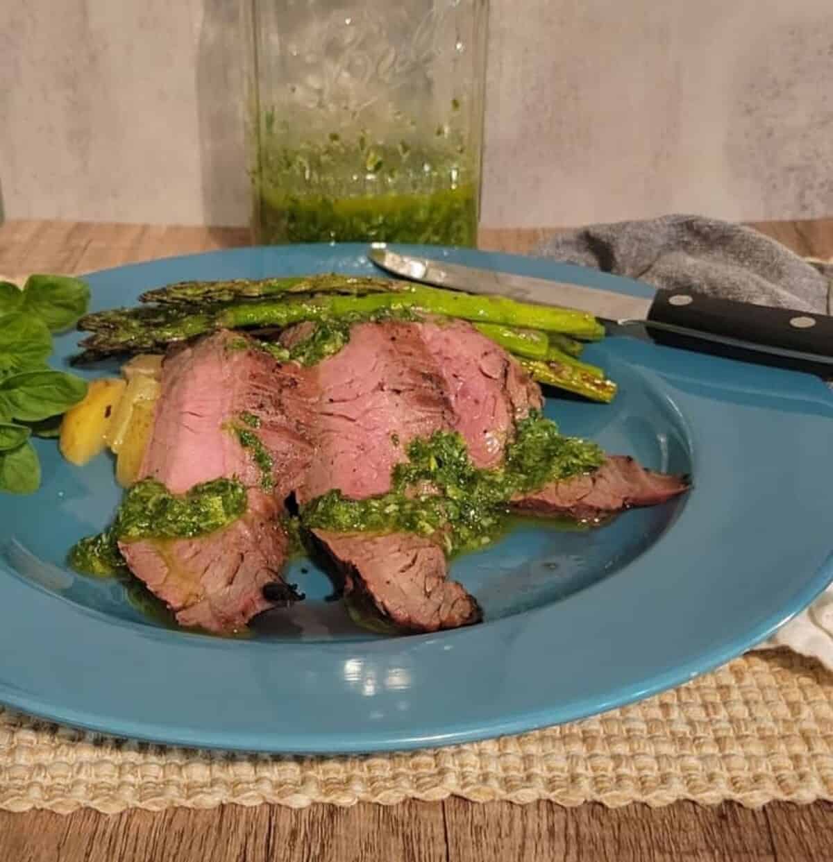 Plated and sliced flank steak with chimichurri, grilled asparagus, roasted potatoes, chimichurri in back ground in mason jar