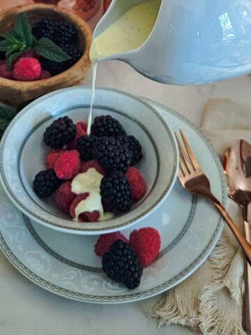 crème anglaise with bourbon sauce being poured over fresh raspberries and blackberries in a bowl next to a wooden bowl with additional fresh berries and fresh mint sprigs