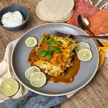 a serving of chicken enchilada casserole on a plate with fresh cilantro and sliced limes, next to a bowl of sour cream and a stack of corn tortillas