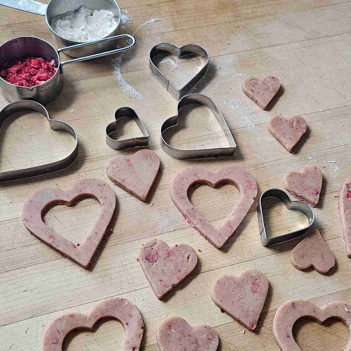 the heart shaped cut out strawberry shortbread cookies on a cutting board, next to heart shaped cookie cutters, flour, and freeze dried strawberries