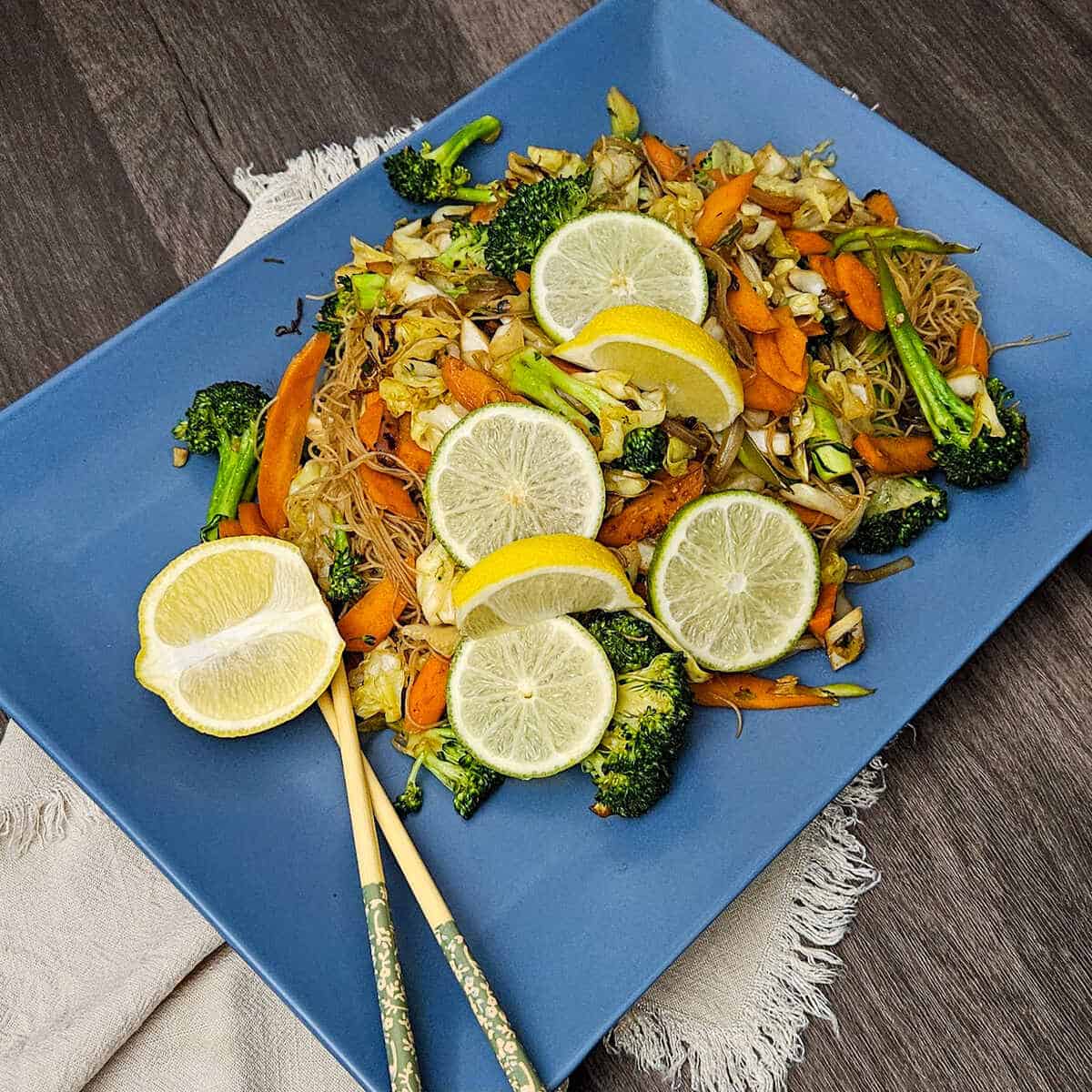 finished easy Asian vegetable stir fry rice noodles on a plate garnished with lemon wedges and lime slices, and chop sticks