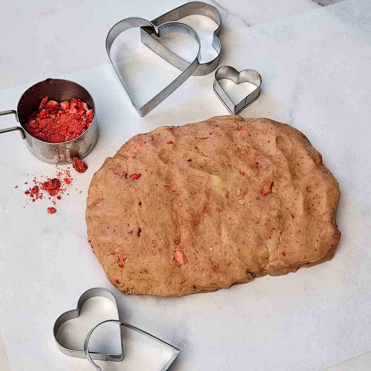 the strawberry shortbread dough finished on parchment paper ready to wrap and refrigerate, next to heart shaped cookie cutters and crushed freeze dried strawberries