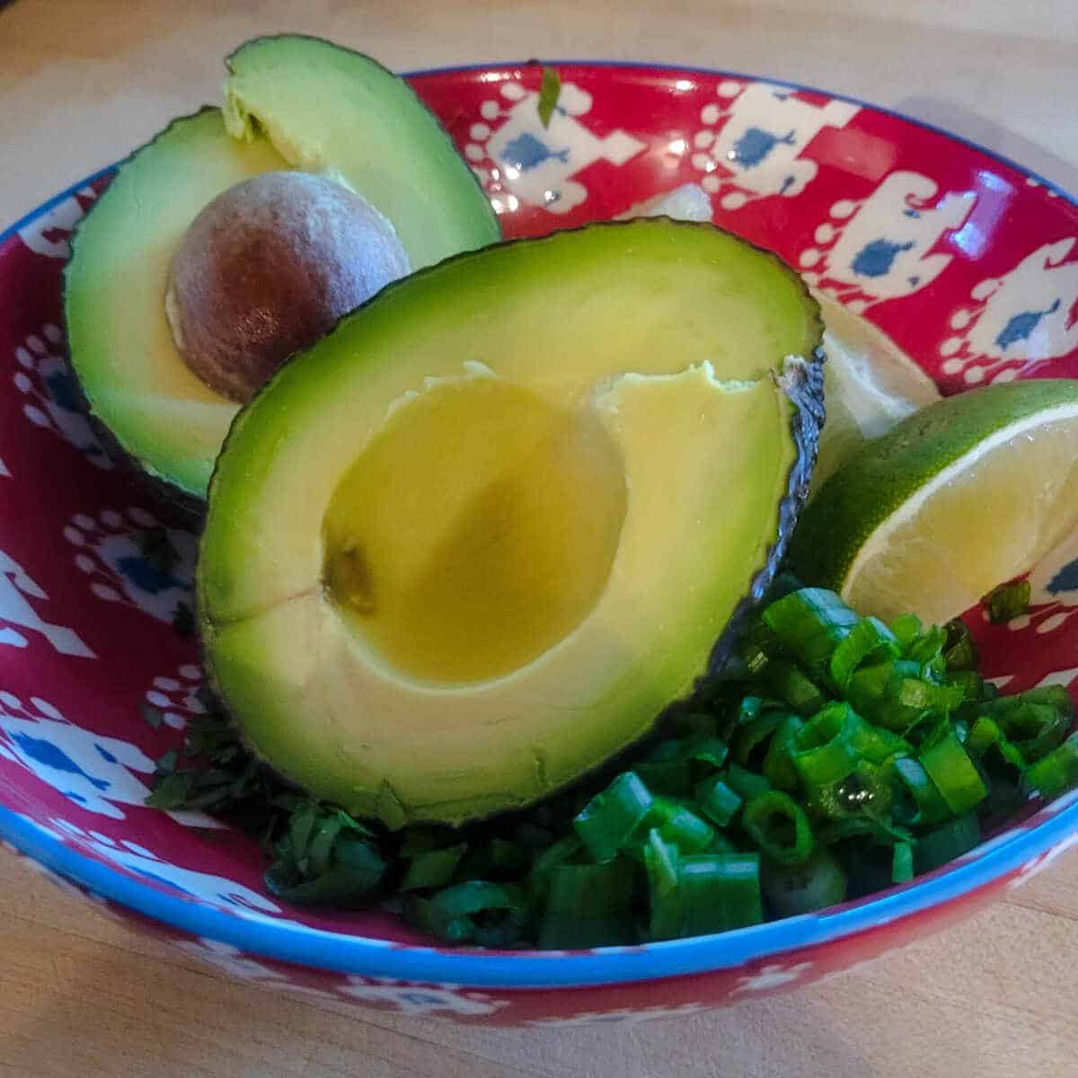 an avocado cut in half, wedges of fresh lime, and sliced green onion in a bowl ready to garnish the chicken enchilada casserole