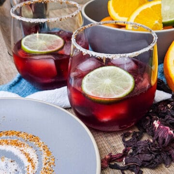 hibiscus margarita mocktail in rimmed glasses garnished with lime slices, next to cut oranges, hibiscus flowers and the dish used to rim the glasses