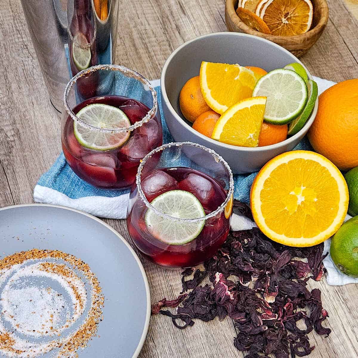 two glasses filled with hibiscus margarita mocktails, garnished with lime wheels, and rimmed with salt and tajin. Oranges and limes, dried hibiscus flowers.