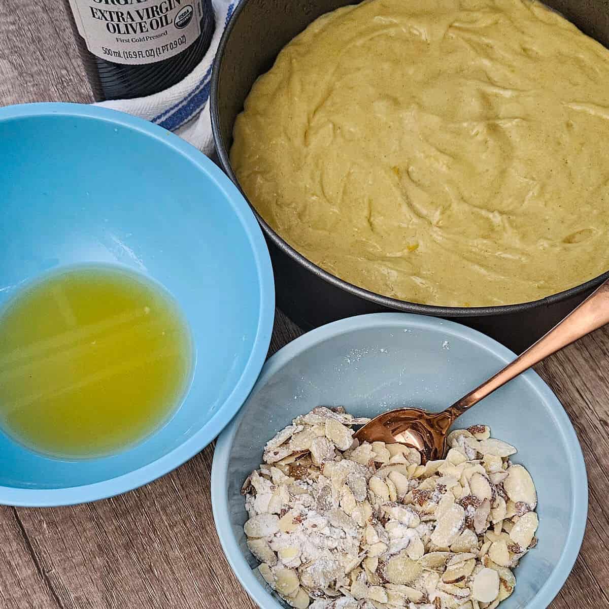 the batter for the gluten free olive oil cake in a pan with bowls containing the almonds and citrus juice for topping