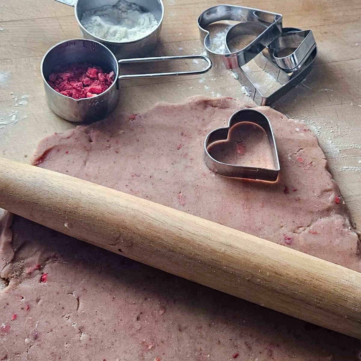 rolling out the strawberry shortbread dough on a cutting board with a wooden rolling pin, next to heart shaped cookie cutters, flour, and freeze dried strawberries
