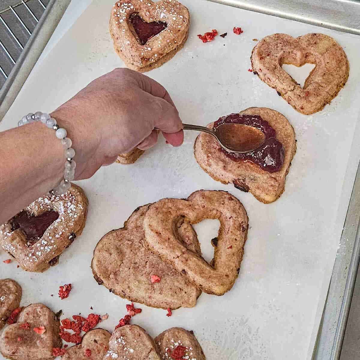spreading jam on the bottom heart shaped strawberry shortbread cookie to create the sandwich with the cut out top cookie