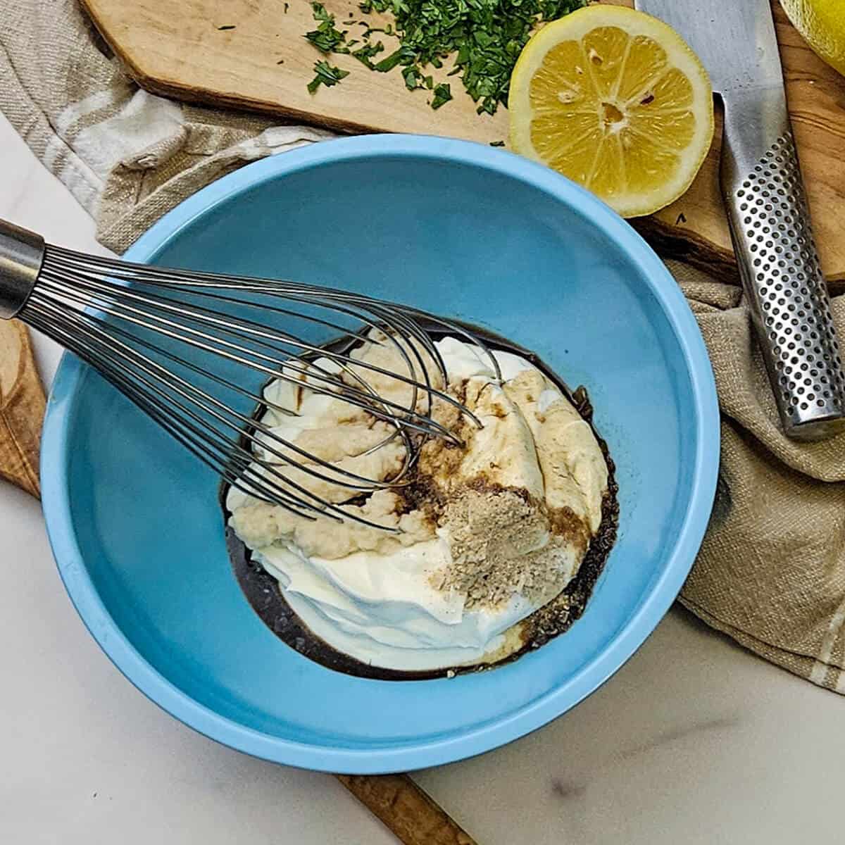 an image of whisking together the ingredients for the best creamy horseradish sauce in a bowl with a wire whisk, with a lemon and chopped parsley