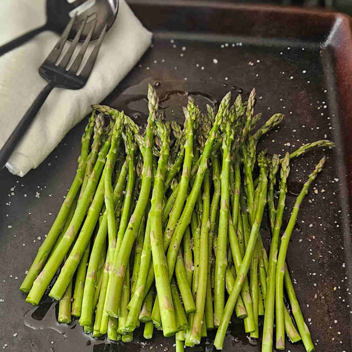 Fresh asparagus on a baking pan seasoned and ready for the oven with a kitchen fork and spoon. For the roasted asparague and fennel salad with golden beets.