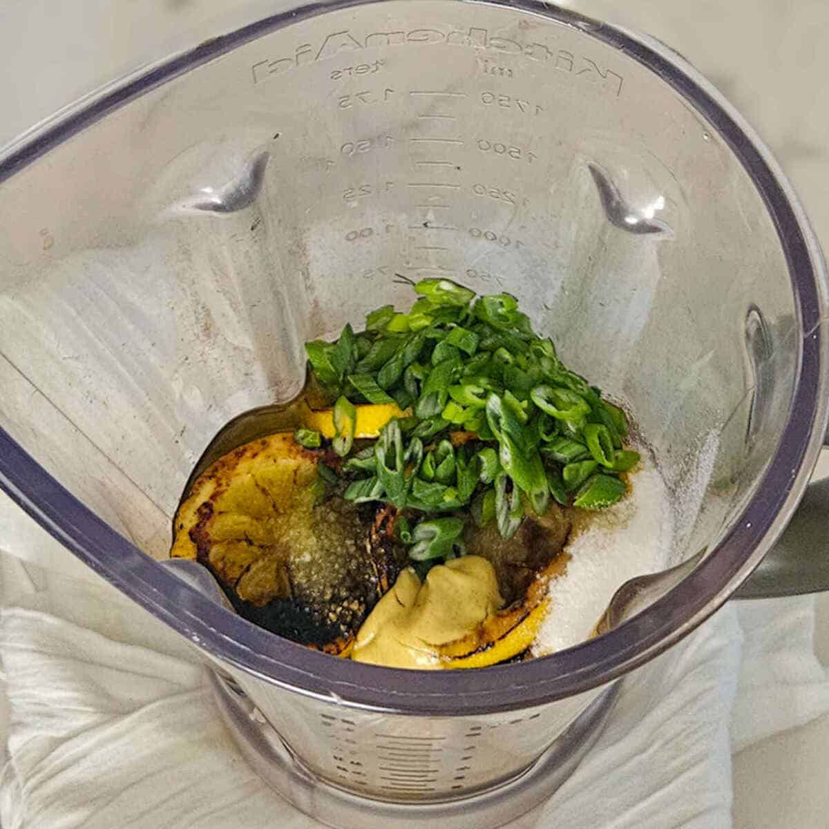 The pitcher of a blender with the whole ingredients for the charred lemon salad dressing recipe, ready to blend together.
