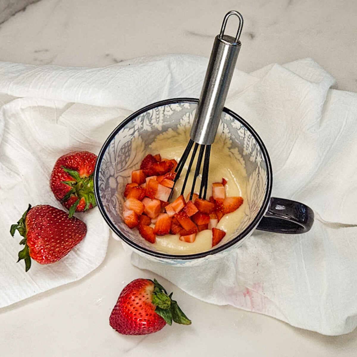 A whisk in a mug with strawberries and batter for the strawberry and cream mug cake.
