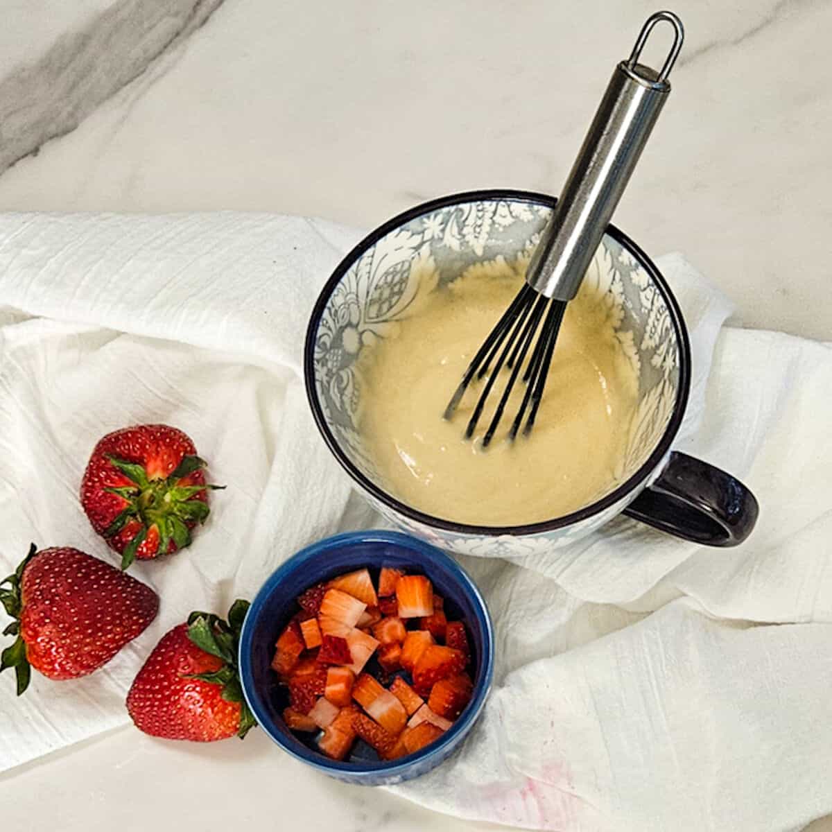 Diced strawberries in a bowl, mug cake batter in a mug with a whisk.