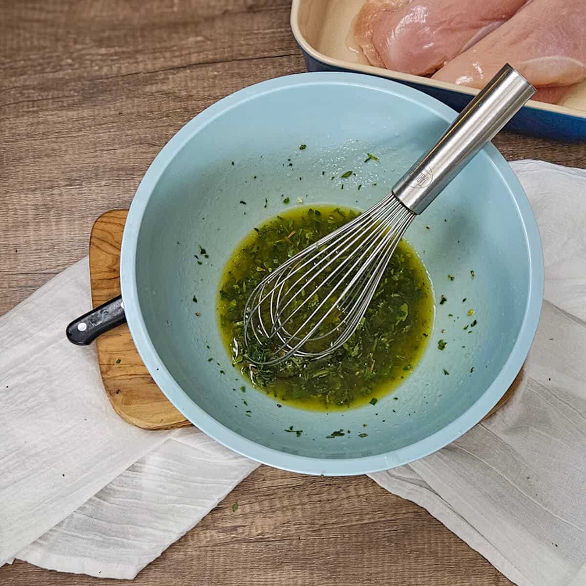 A bowl with white balsamic herb marinade and a whisk on a cutting board.