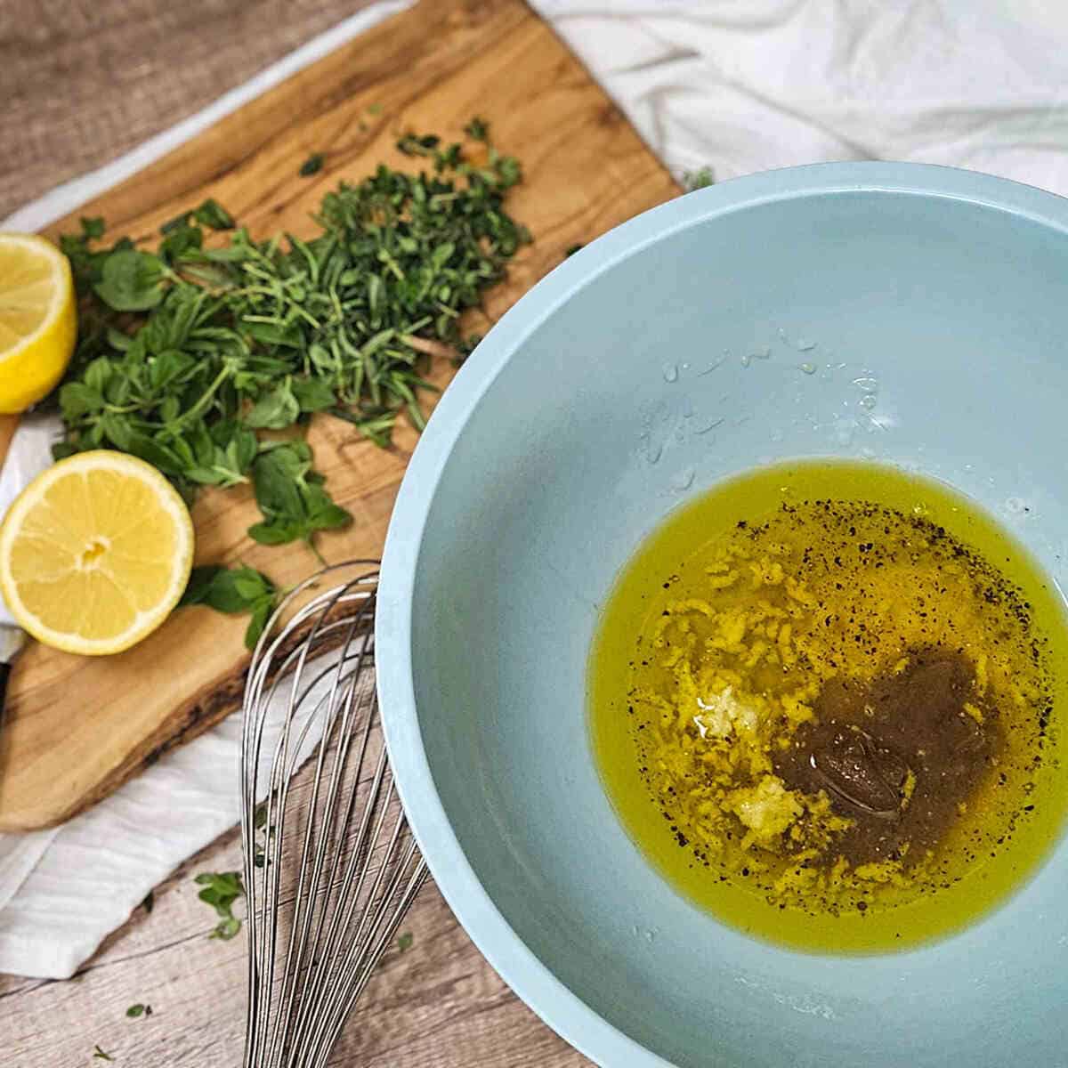 A bowl of olive oil and ingredients for white balsamic herb marinade, herbs and lemon halves on a cutting board.
